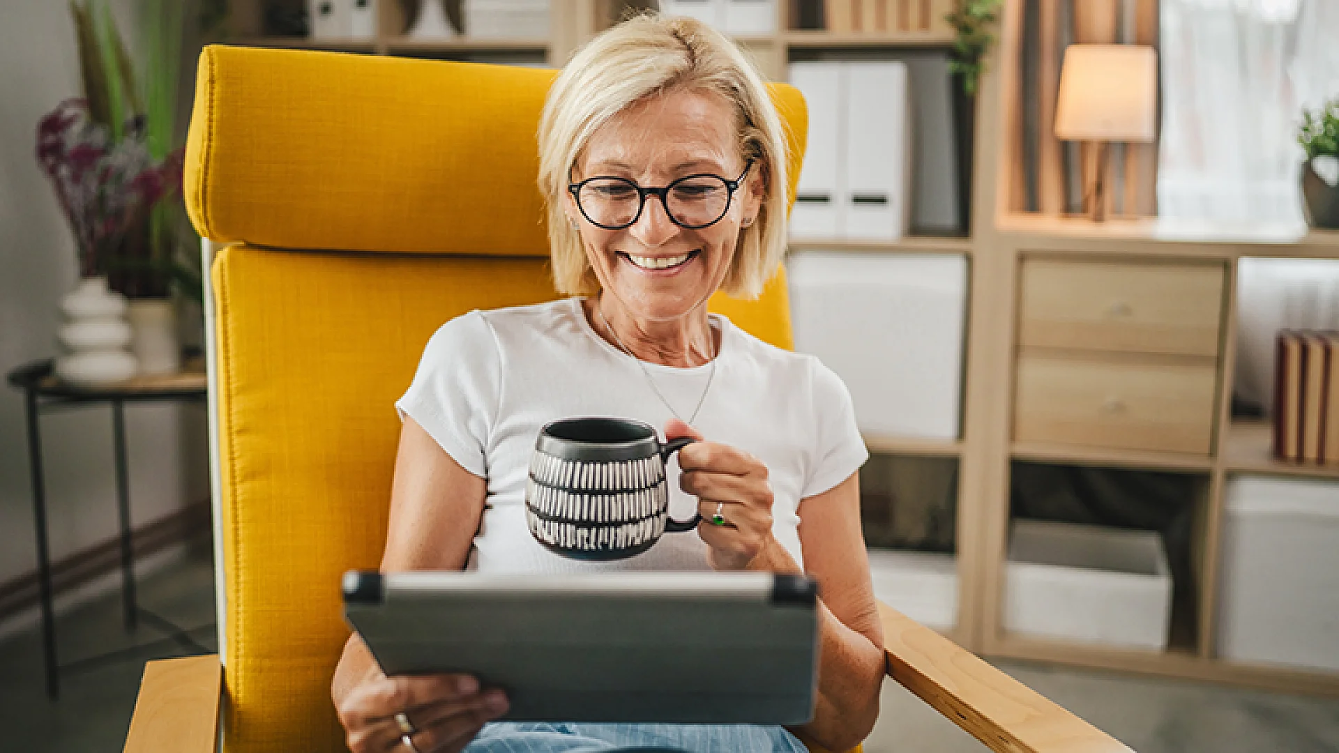 Lächelnde Frau liest entspannt auf einem Tablet und trinkt Kaffee im Sessel