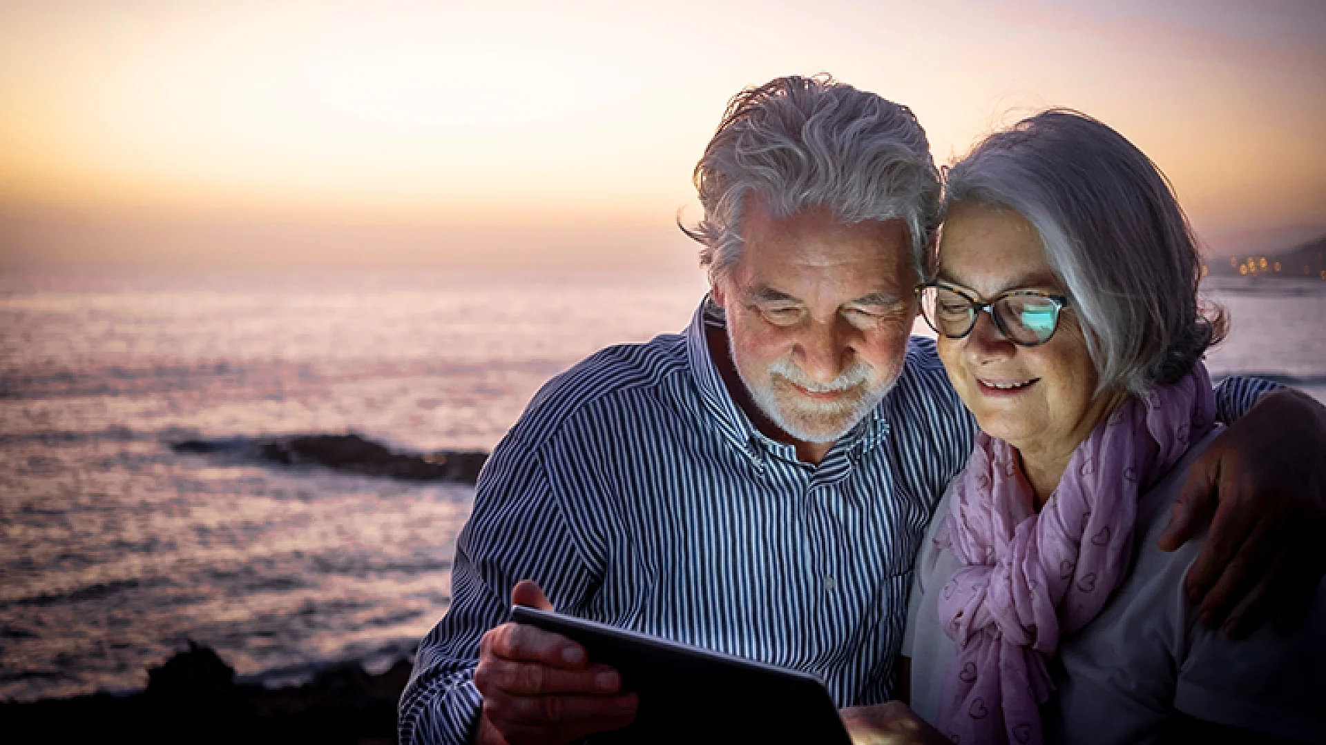 Paar liest gemeinsam auf einem Tablet am Meer bei Sonnenuntergang