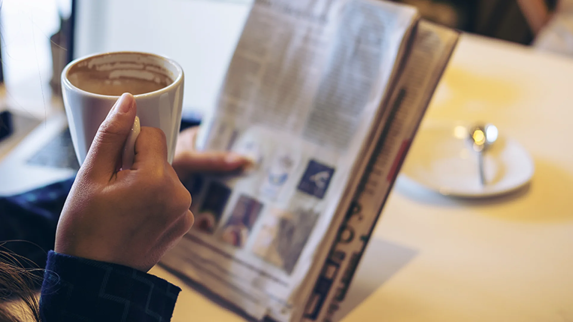 Person liest bei einer Tasse Kaffee entspannt eine gedruckte Zeitung am Tisch