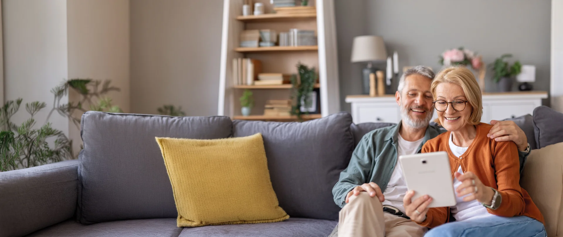 Lachendes älteres Paar sitzt gemeinsam auf Sofa und hält ein Tablet in modernem Wohnzimmer